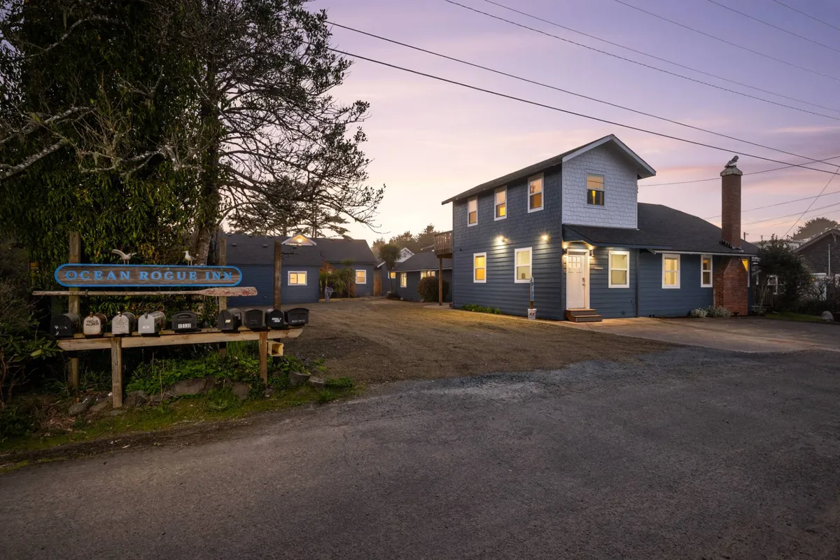 This image features the Ocean Rogue Inn sign in front of the blue building, surrounded by lush trees, with a warmly lit entrance that feels inviting. The setting hints at communal spaces where guests can relax, gather, or enjoy amenities like a fire pit after a day spent outdoors.