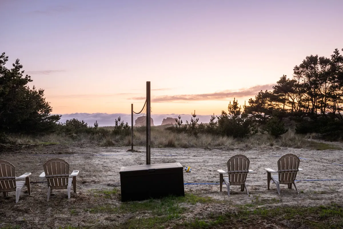 The image features Adirondack chairs arranged near a volleyball net, with distant rock formations and a glowing sunset sky as the backdrop. The setting suggests a space where guests can relax after activities, enjoy the sunset, and mingle with others while appreciating the coastal view.