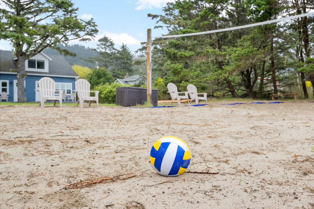 A cheerful scene featuring a volleyball net set up in a sandy area, surrounded by adirondack chairs and lush trees. The setup invites energetic play, relaxation, and socializing, capturing the lively spirit of vacation fun for families and small groups.