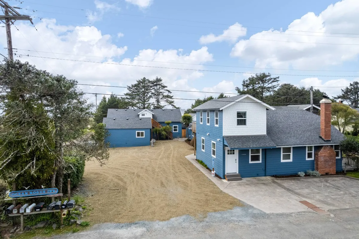 A view of the blue buildings of the inn, a gravel driveway, and a signpost, with outdoor spaces suited for gatherings or barbecues. The mood is inviting, with hints of sunset and plenty of space for guests to mingle, stargaze, or enjoy evening fire-pit gatherings.