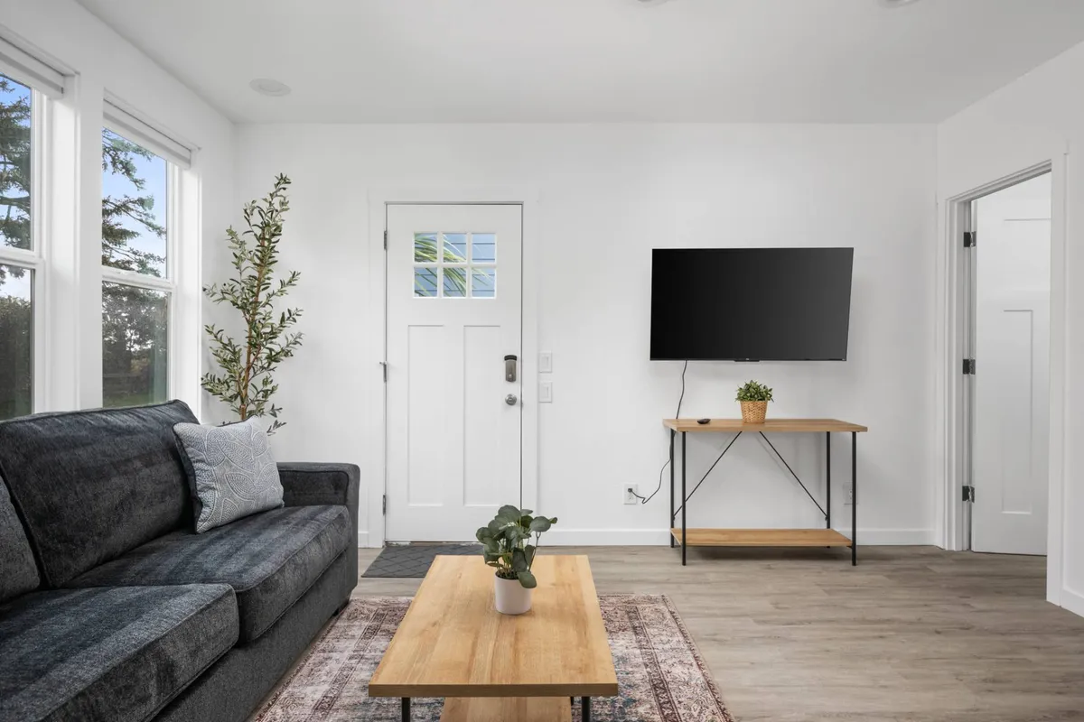A cozy, well-lit living area featuring a comfortable sofa, a coffee table, a television, and a doorway leading to other parts of the unit. There's also a potted plant adding a touch of greenery. The open layout and new furnishings are visible, creating an inviting space for relaxation.