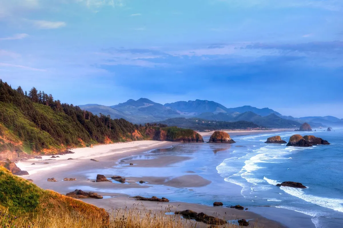 A sweeping view of the sandy beach with the majestic Twin Rocks Arch rising from the ocean, gentle waves rolling in, and distant mountains on the horizon. The tranquil scene includes scattered seashells and hints at beachcombing, hiking, and relaxation activities.