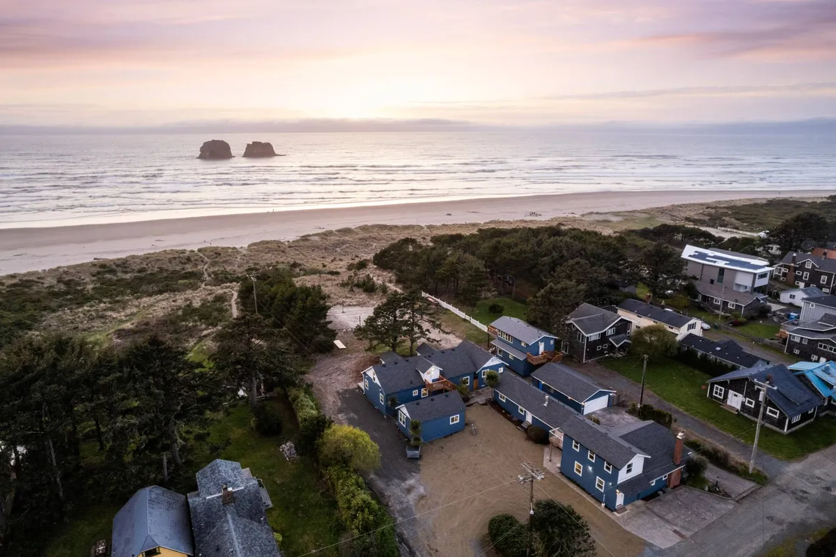 A serene beach scene featuring soft sand, distinctive sea stacks in the distance, and nearby houses and trees. The scene suggests easy access to the shore for activities like beachcombing, sunset watching, and sandcastle building.