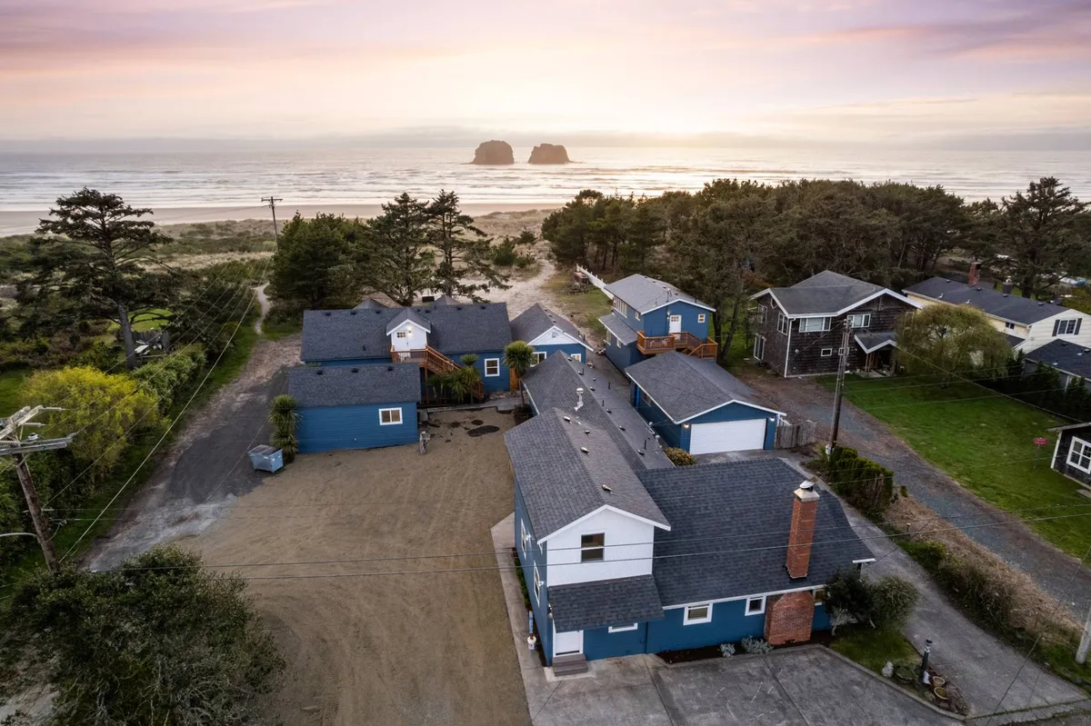 A sweeping view of the ocean framed by unique rock formations in the distance, with a cluster of houses visible in the foreground. The arrangement suggests a community setting right on the beach, with plenty of open space for gathering and outdoor activities. Subtle hints of a fire pit gathering area add to the communal, family-friendly vibe.