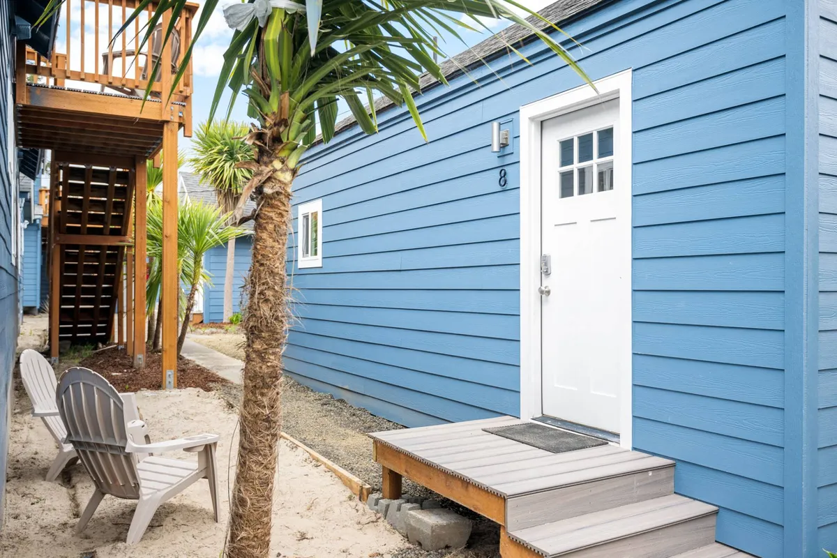 A welcoming exterior view of the Ocean Rogue Inn, featuring its blue facade, white door, and wooden steps. Adirondack chairs are arranged outside, beneath a palm tree, suggesting a casual spot for guests to gather, relax, and enjoy the outdoors. The ambient lighting hints at sunset, creating a warm, social atmosphere.