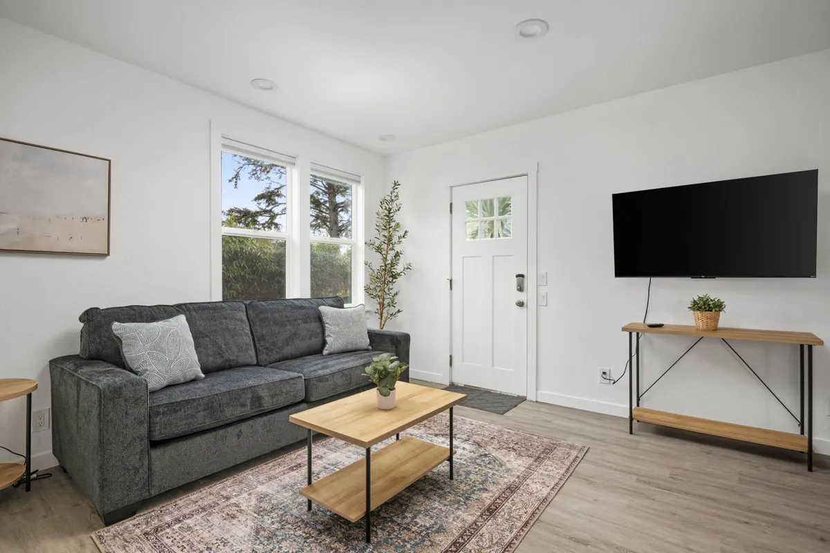 The image showcases a cozy condo living room with a comfortable sofa, coffee table, television, front door, and lush plants. Soft morning light filters through a nearby window, creating an inviting space perfect for relaxing, enjoying coffee, or gathering for a movie night with friends.