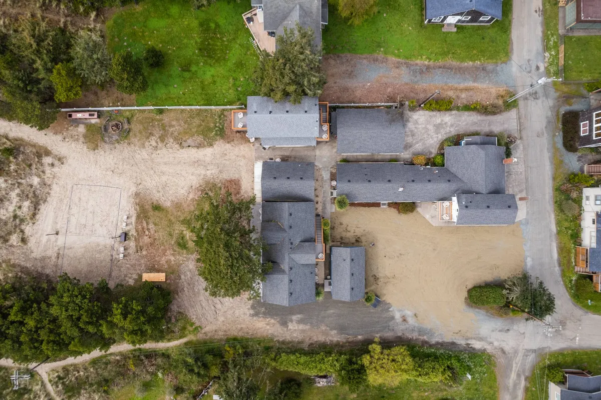 An open sandy area bordered by a cluster of buildings and lush greenery, ideal for outdoor games or group activities. The space is clearly designed for communal use, implying volleyball matches, summer barbecues, or spontaneous gatherings—a hub for vibrant social interaction.