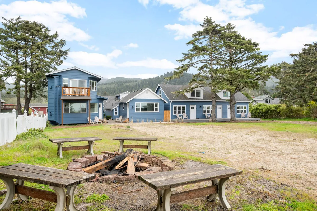 A communal fire pit is surrounded by blue houses and large trees, evoking warmth and togetherness. The fire pit area is clearly set up for group gatherings, with ample space for guests to share stories, roast marshmallows, and enjoy evenings under the stars.